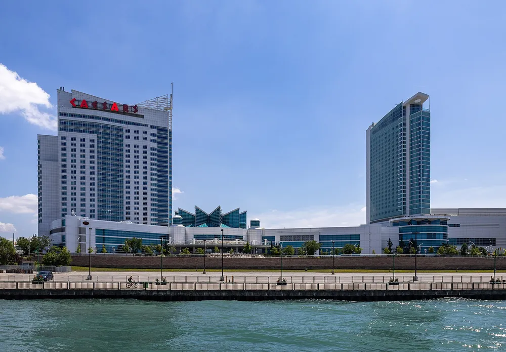 Photograph: the two hotel towers and the low casino building of Caesars Windsor seen from the Detroit River on a sunny day, with the riverside walkway in front.